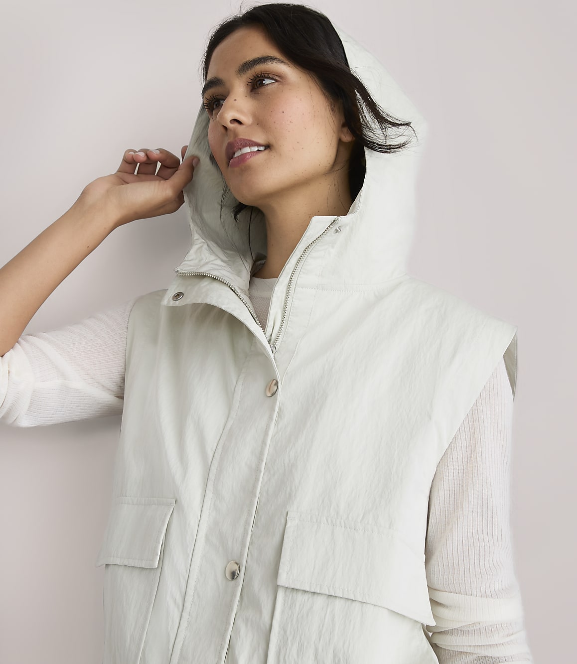 A woman in a light-colored hooded vest and green pants stands against a plain white background.