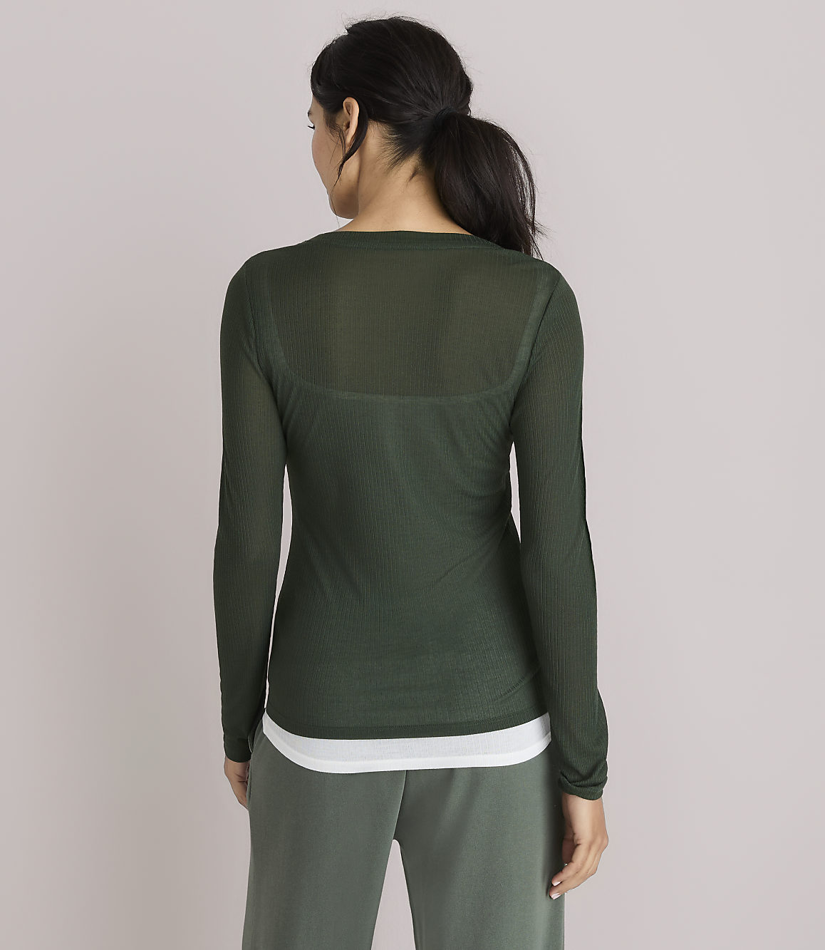 A woman in a sheer green long-sleeved shirt and white tank top stands against a plain, light gray background.