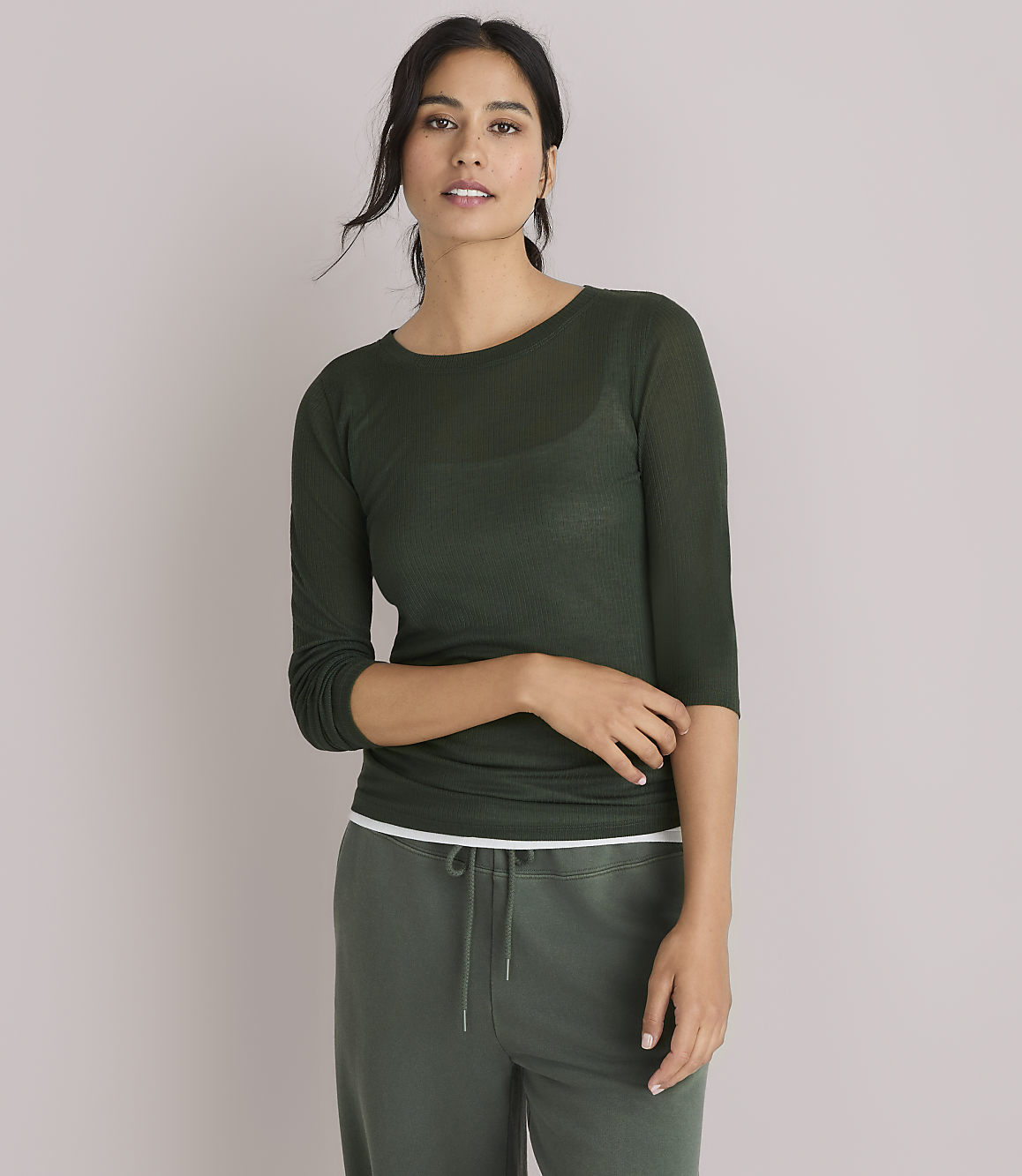 A woman in a dark green, sheer, long-sleeved shirt and green pants poses against a plain white background.