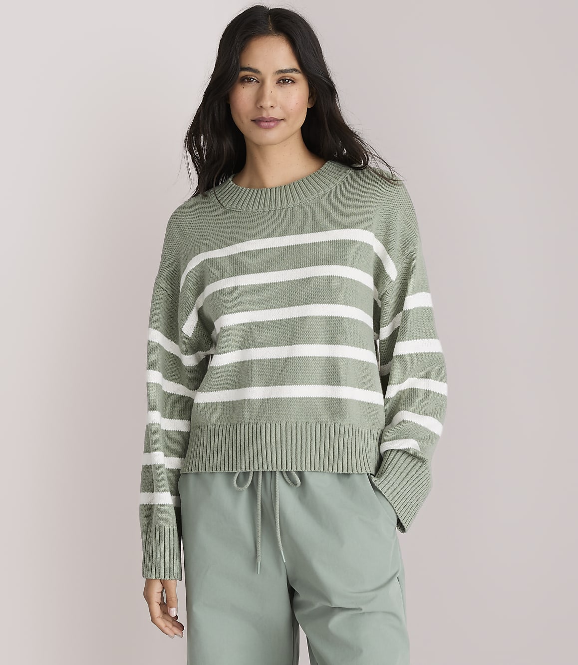 A woman in a green and white striped sweater and light green pants stands against a plain gray background.