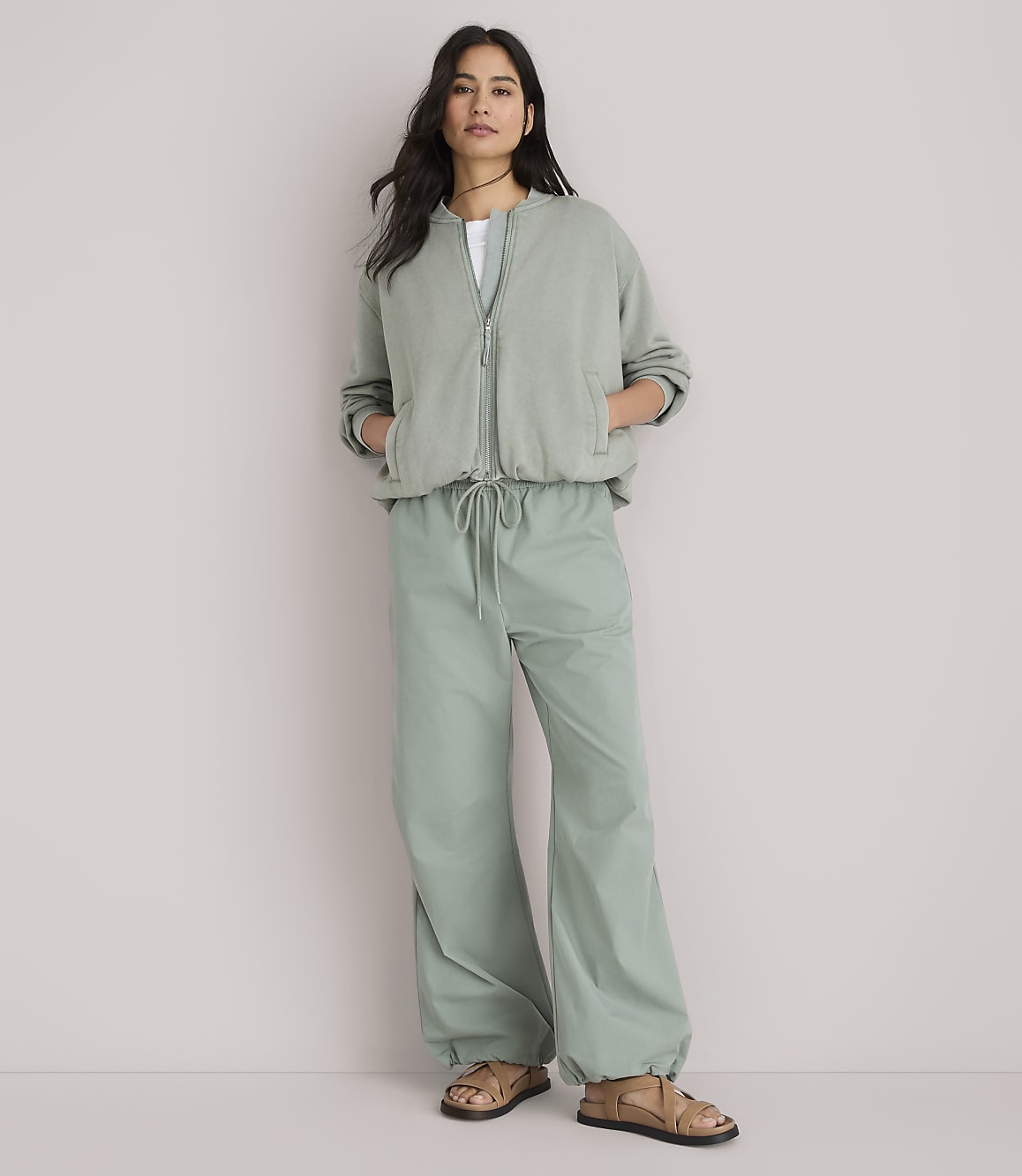 A woman stands in the foreground wearing a sage green bomber jacket and matching wide-leg pants, against a plain white background.