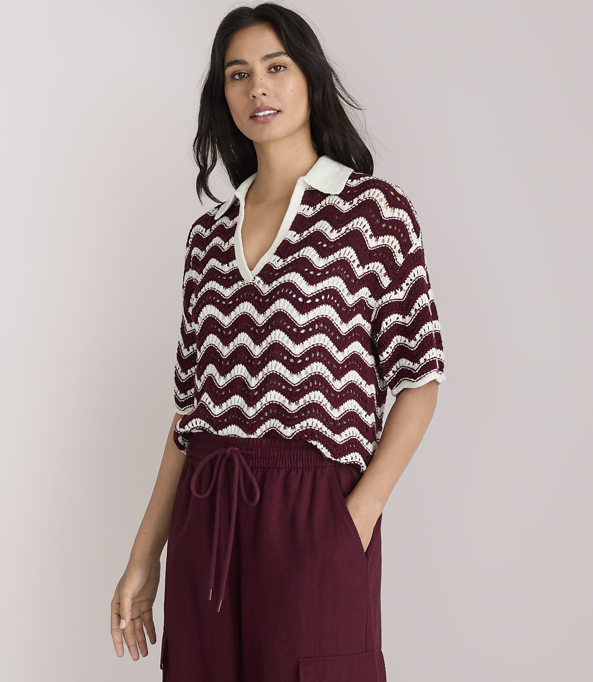 A woman in a chevron-patterned top and maroon wide-leg pants stands against a plain white background.