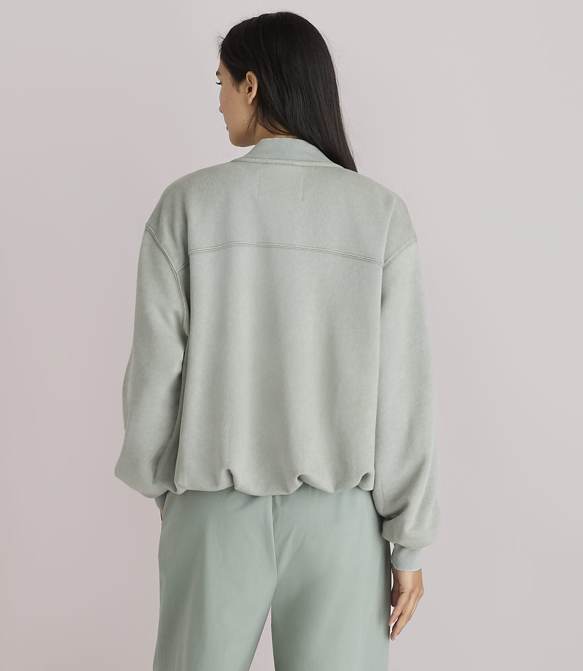 A woman in a light green sweatshirt and pants stands against a plain white background.