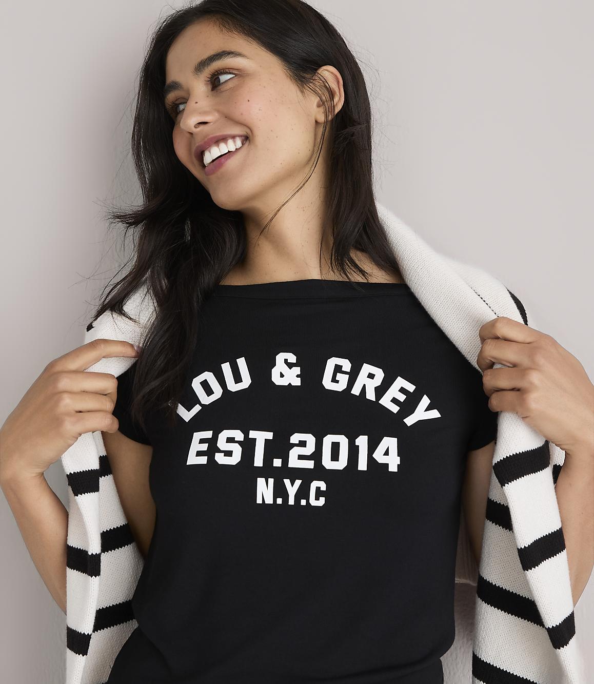 A smiling woman in a black t-shirt with "LOU & GREY EST. 2014 N.Y.C." printed on it, draped with a striped scarf, stands against a plain white background.