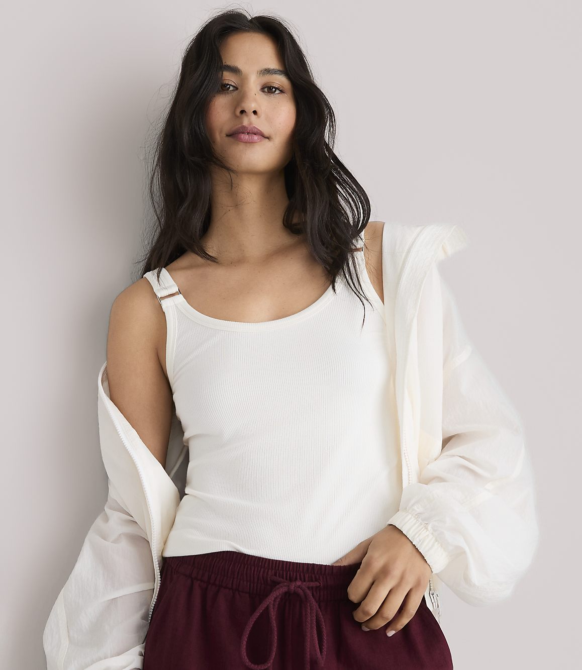 A woman in a white tank top and maroon pants stands against a plain white background.