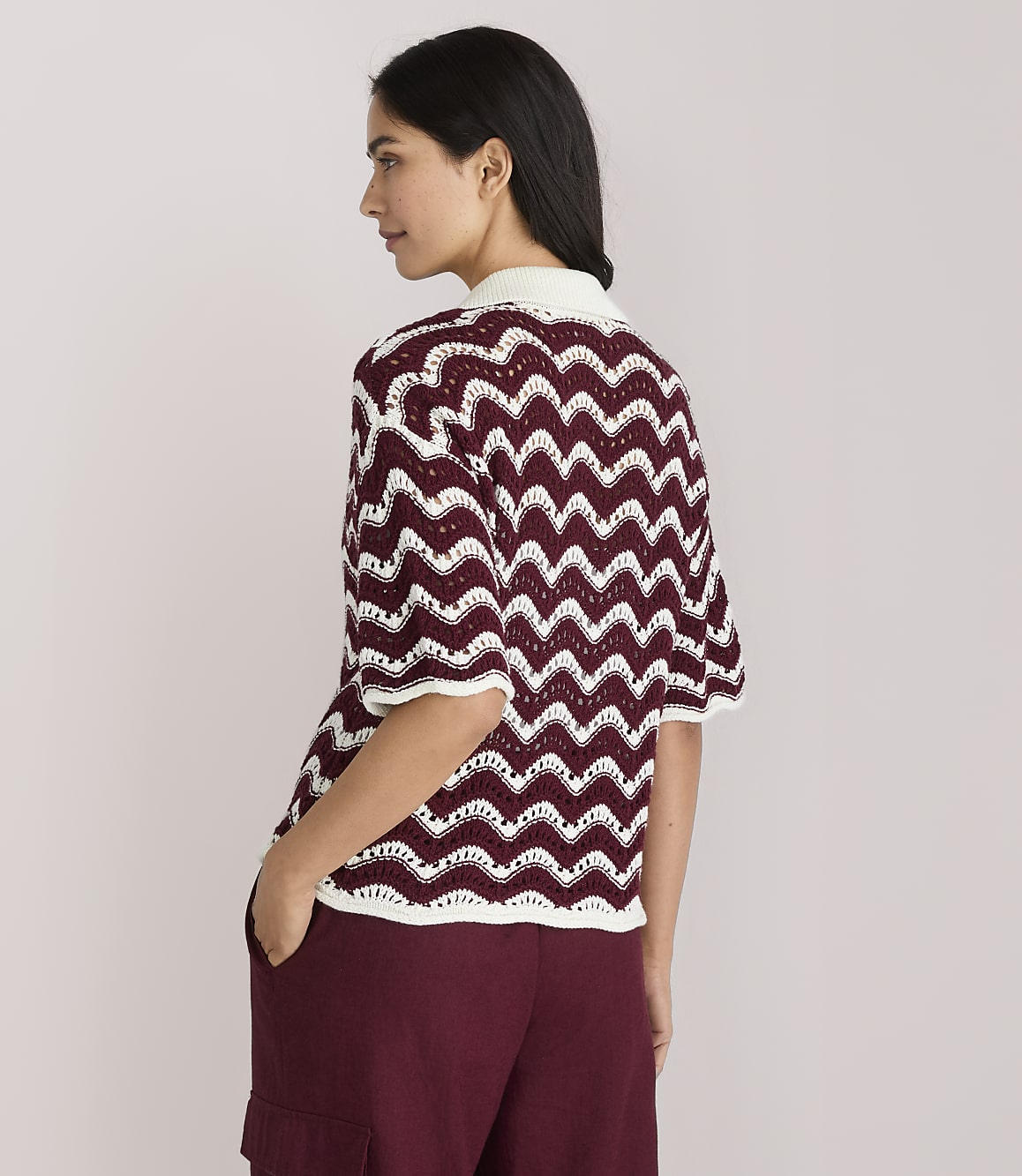 A woman in a burgundy and white chevron knit top and burgundy pants stands with her back to the viewer against a plain white background.