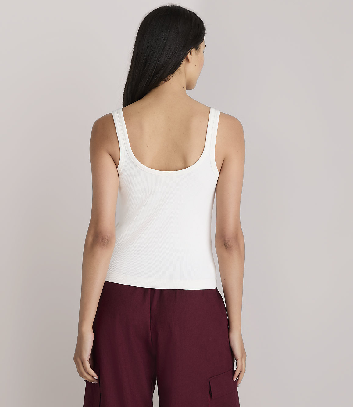 A woman in a white tank top and maroon pants stands against a plain white background.