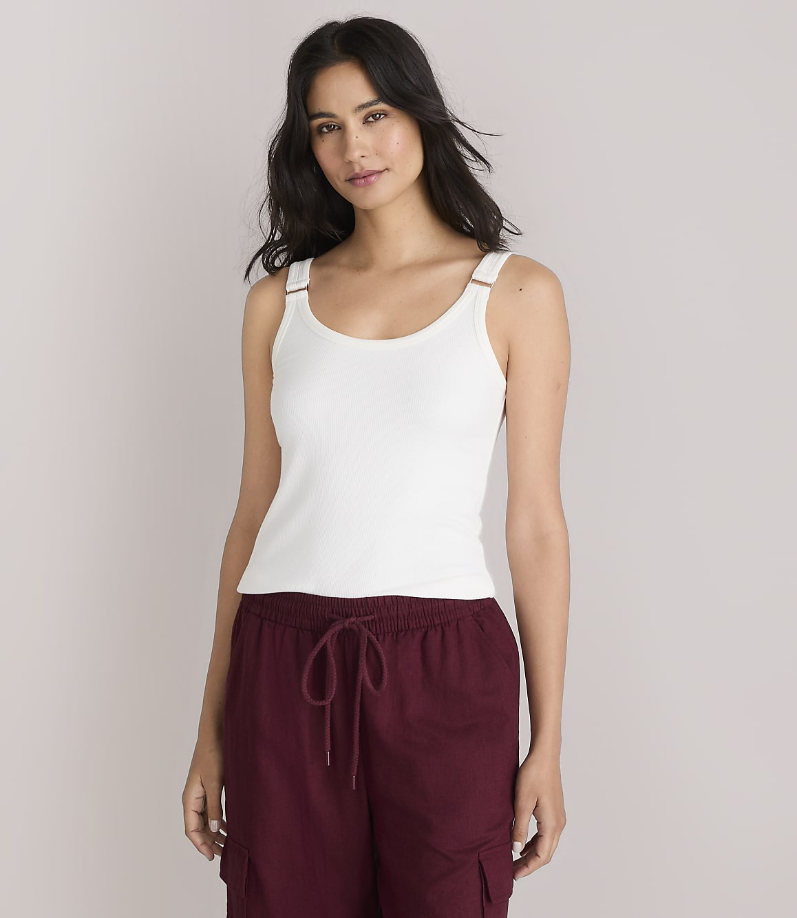A woman in a white tank top and maroon pants stands against a plain white background.