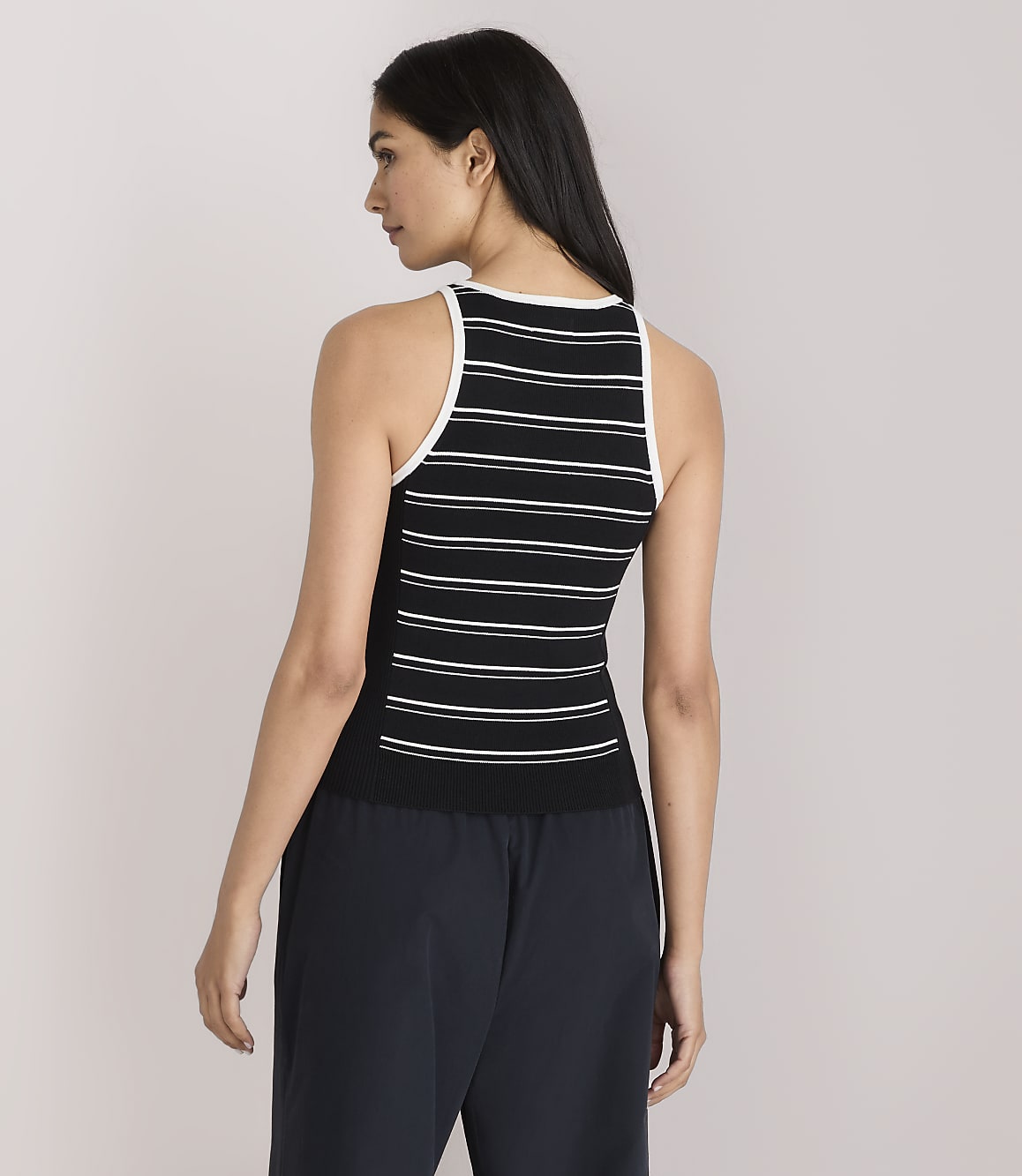 A woman in a black and white striped tank top and dark pants stands against a plain white background.