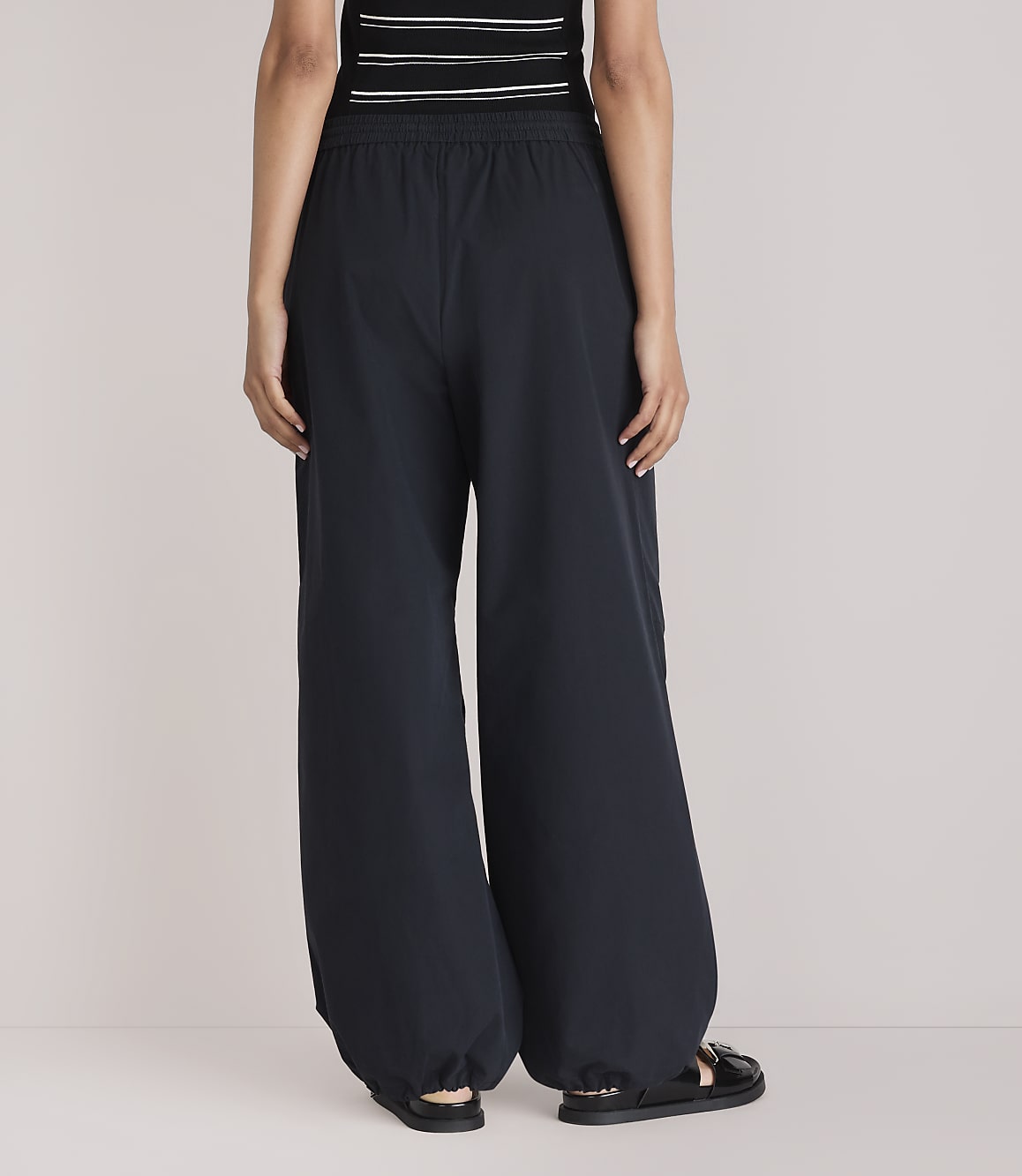 A person wearing dark, wide-leg pants and sandals stands against a plain white background.