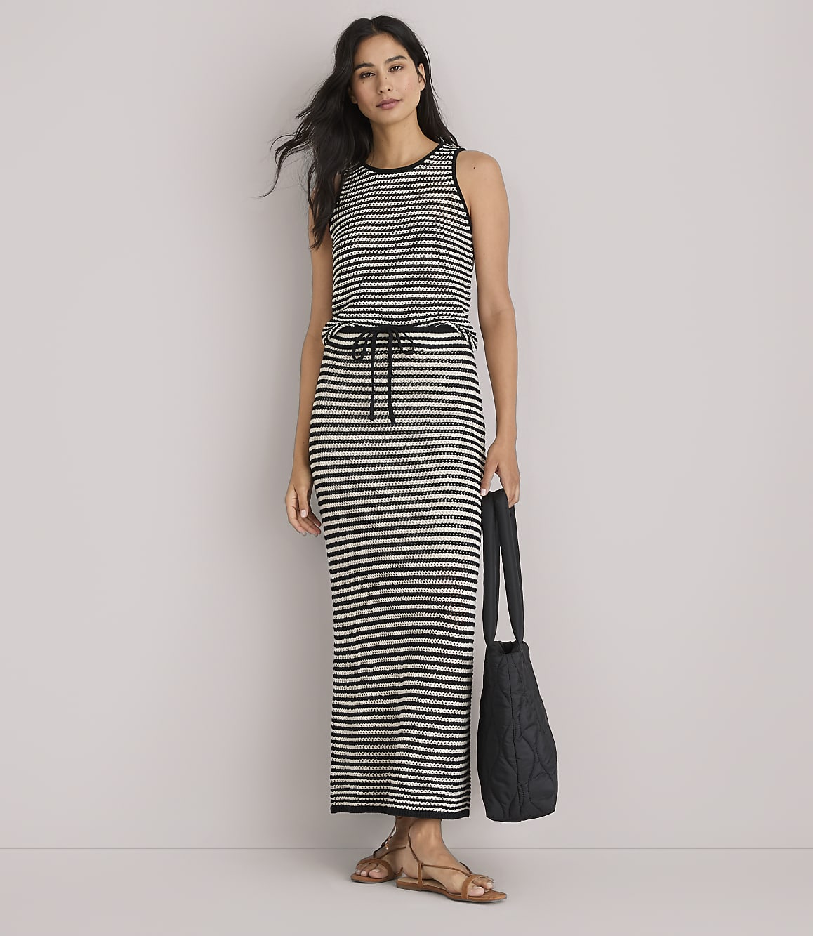 A woman in a striped maxi dress and sandals stands against a plain white background, holding a black quilted tote bag.