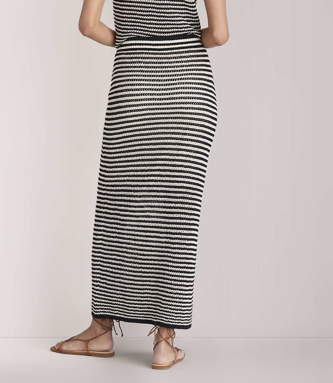 A person wearing a black and white striped maxi skirt and brown sandals stands against a plain white background.