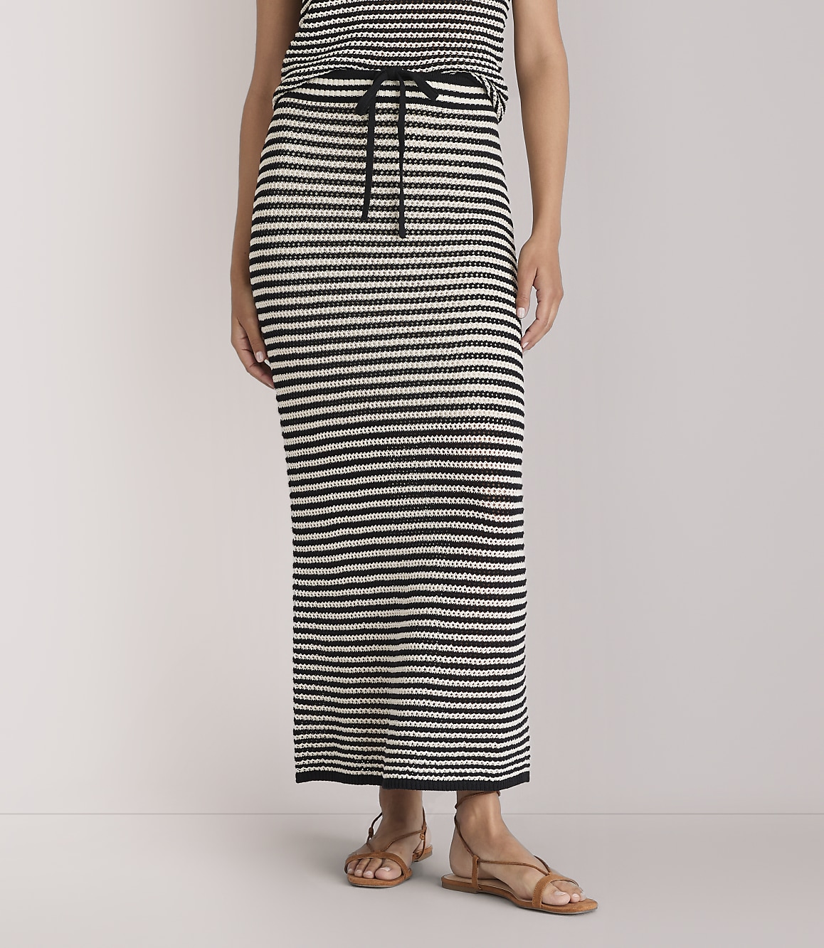 A person stands wearing a black and white striped maxi skirt and sandals against a plain white background.