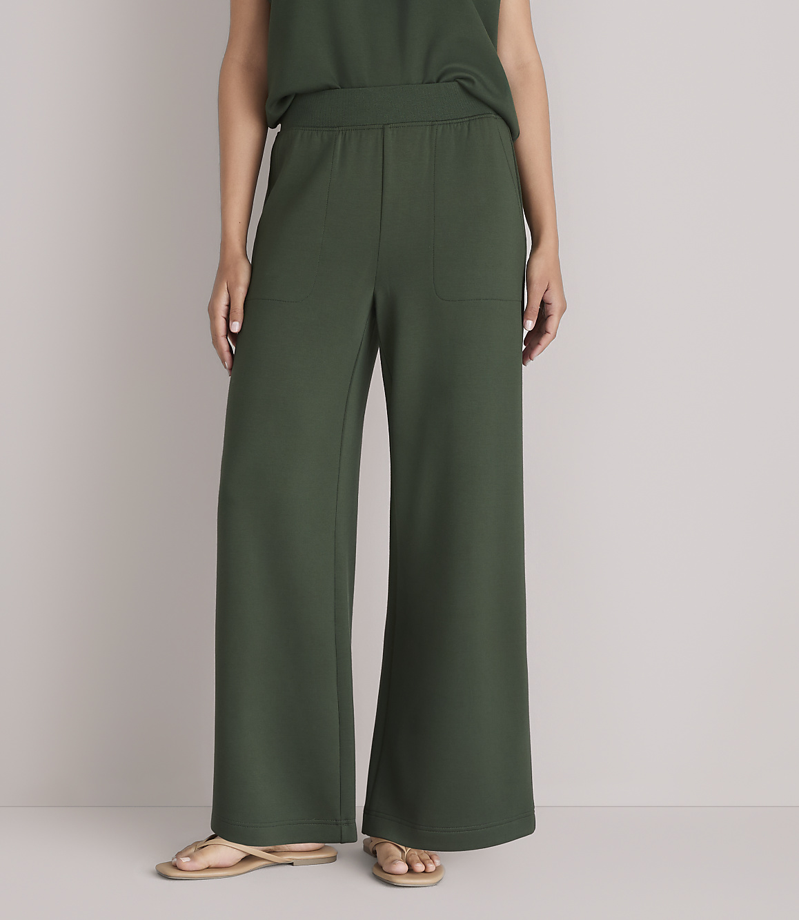 A person wearing dark green wide-leg pants and sandals stands against a plain white background.