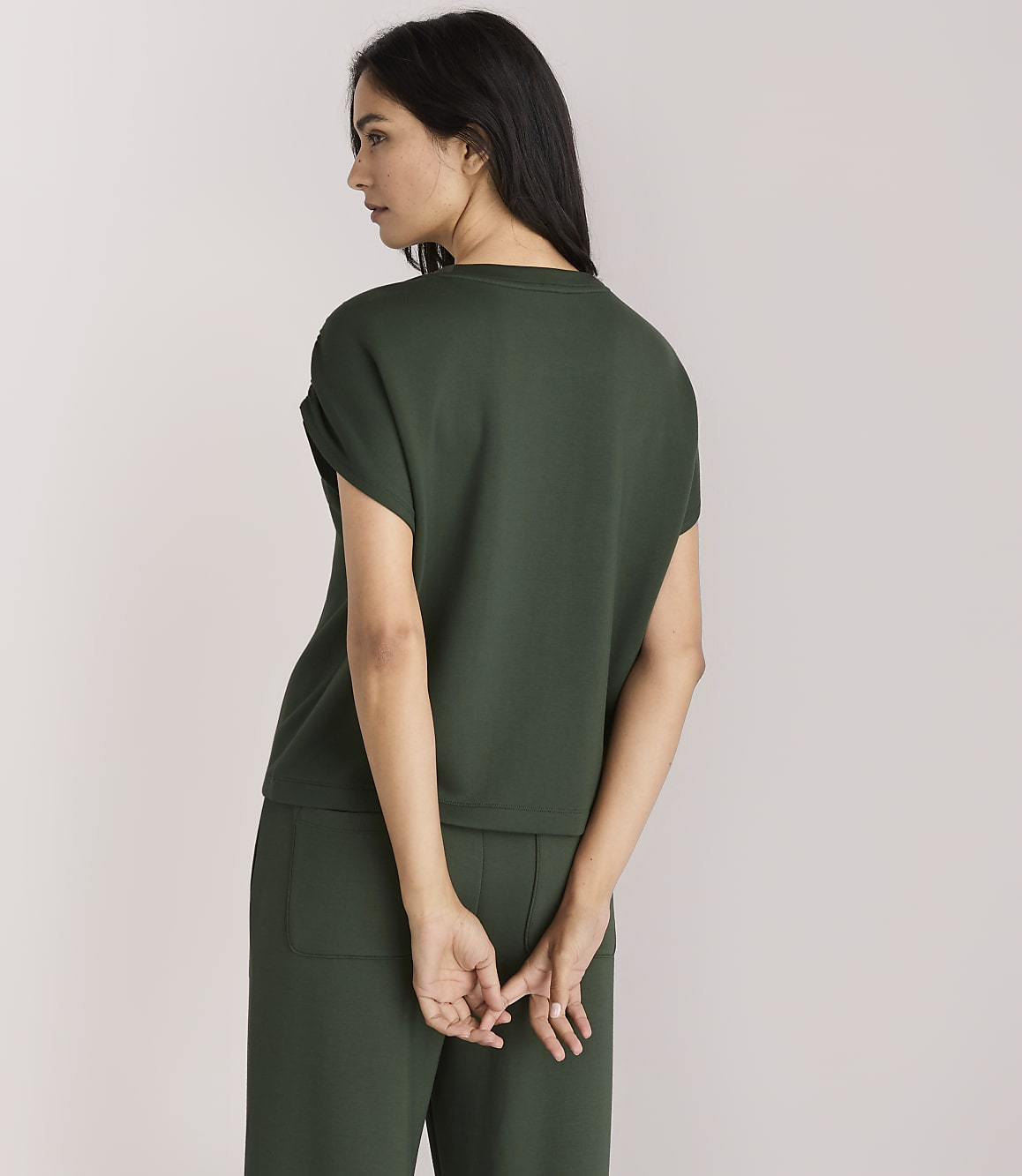A woman in a dark green top and pants stands with her back to the viewer against a plain white background.