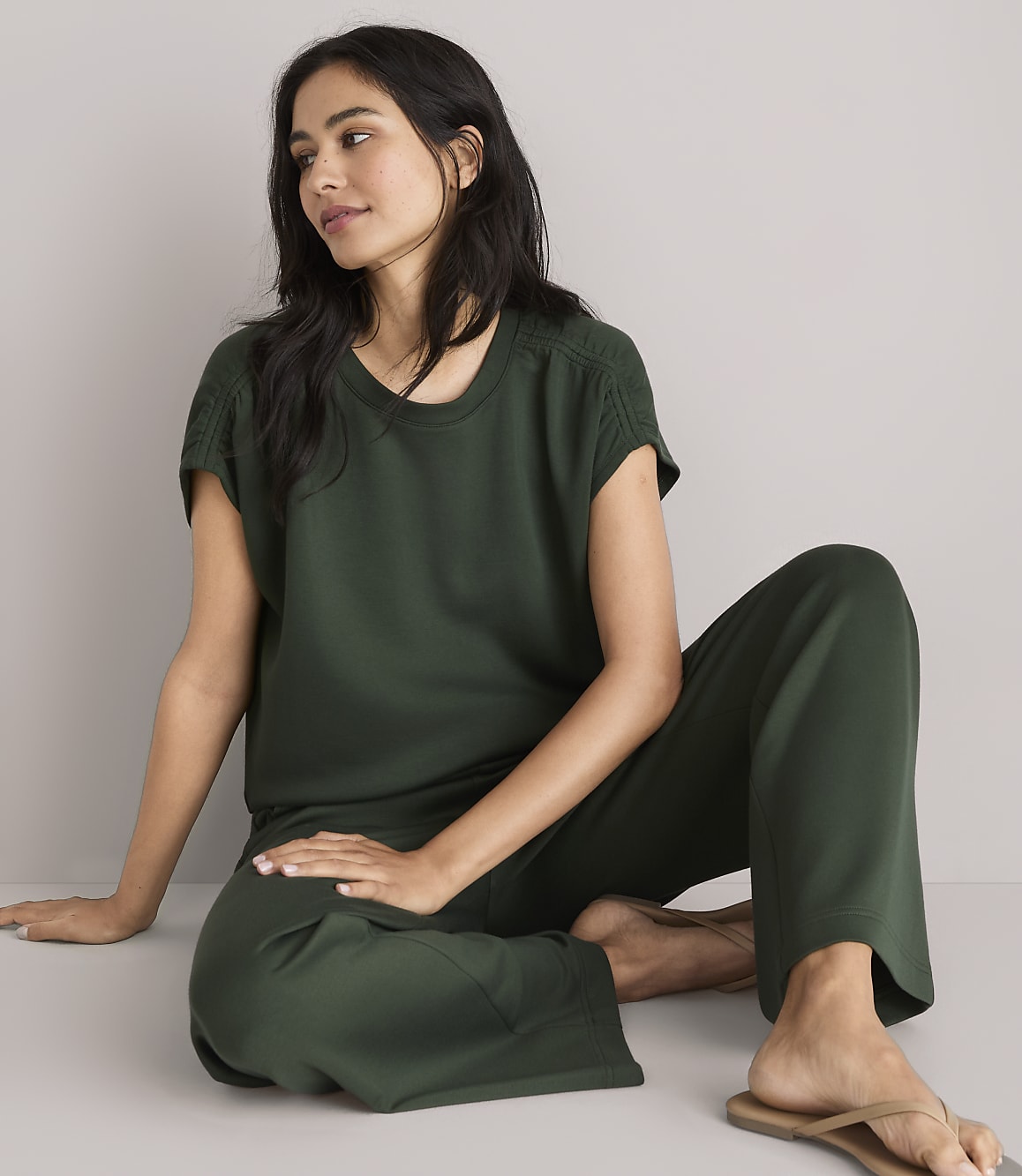 A woman in a dark green loungewear set sits on a white floor, looking to the side with a plain white background.