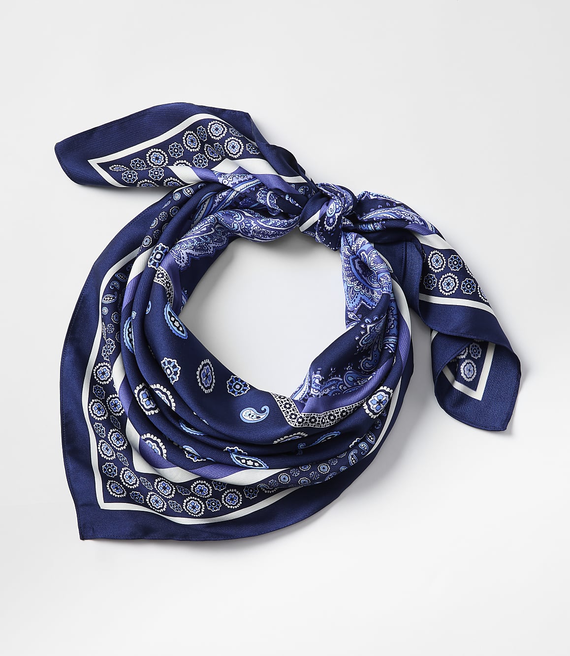 A blue paisley patterned silk scarf is tied in a knot in the foreground against a plain white background.