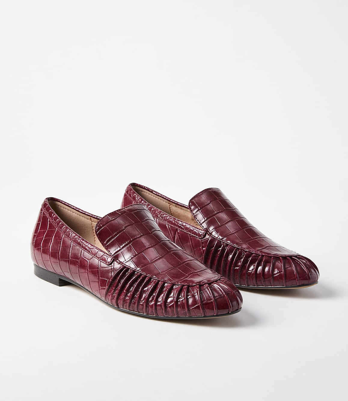 A pair of burgundy croc-embossed leather loafers with pleated detailing sits on a plain white background.