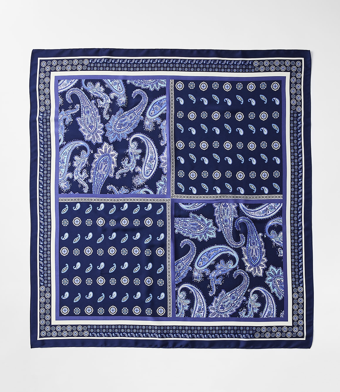 A square silk scarf with a dark blue border features four distinct quadrants of paisley and floral patterns in shades of blue and white, set against a plain white background.
