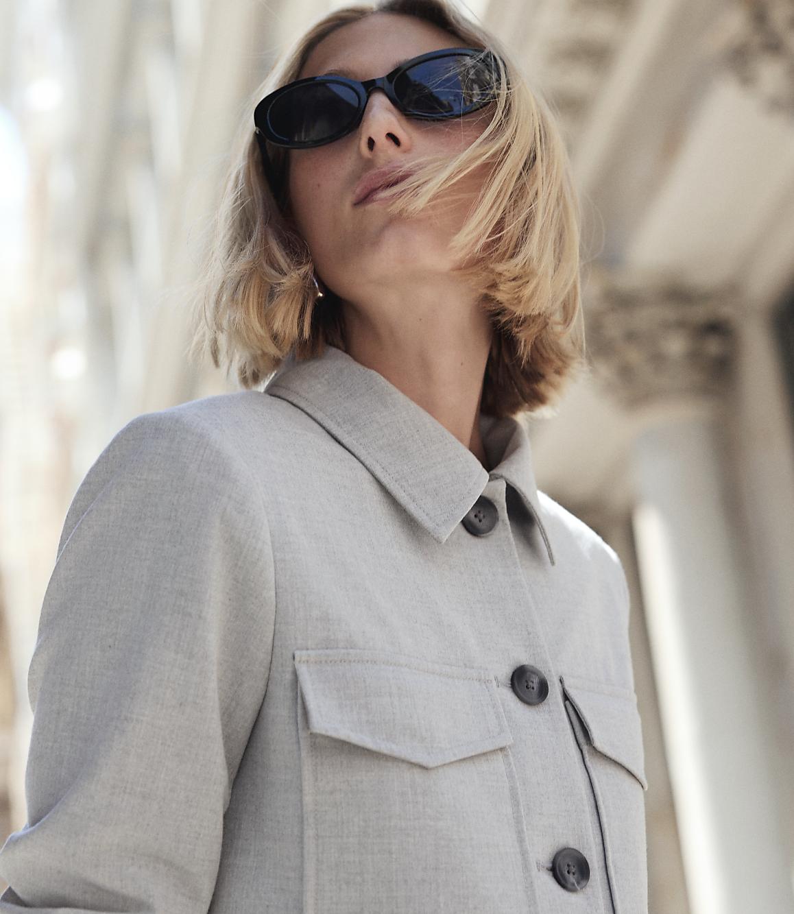A woman in sunglasses and a light-colored jacket stands in front of a blurred architectural background.