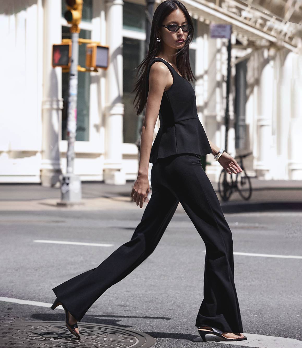 A woman in a black peplum top and flared pants walks across a street with a traffic light and buildings in the background.
