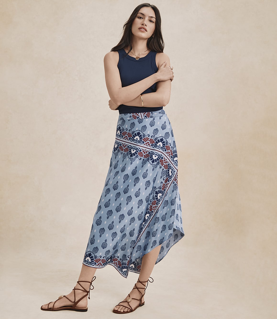 A woman in a blue tank top and a flowing, patterned skirt stands against a plain, light brown background.