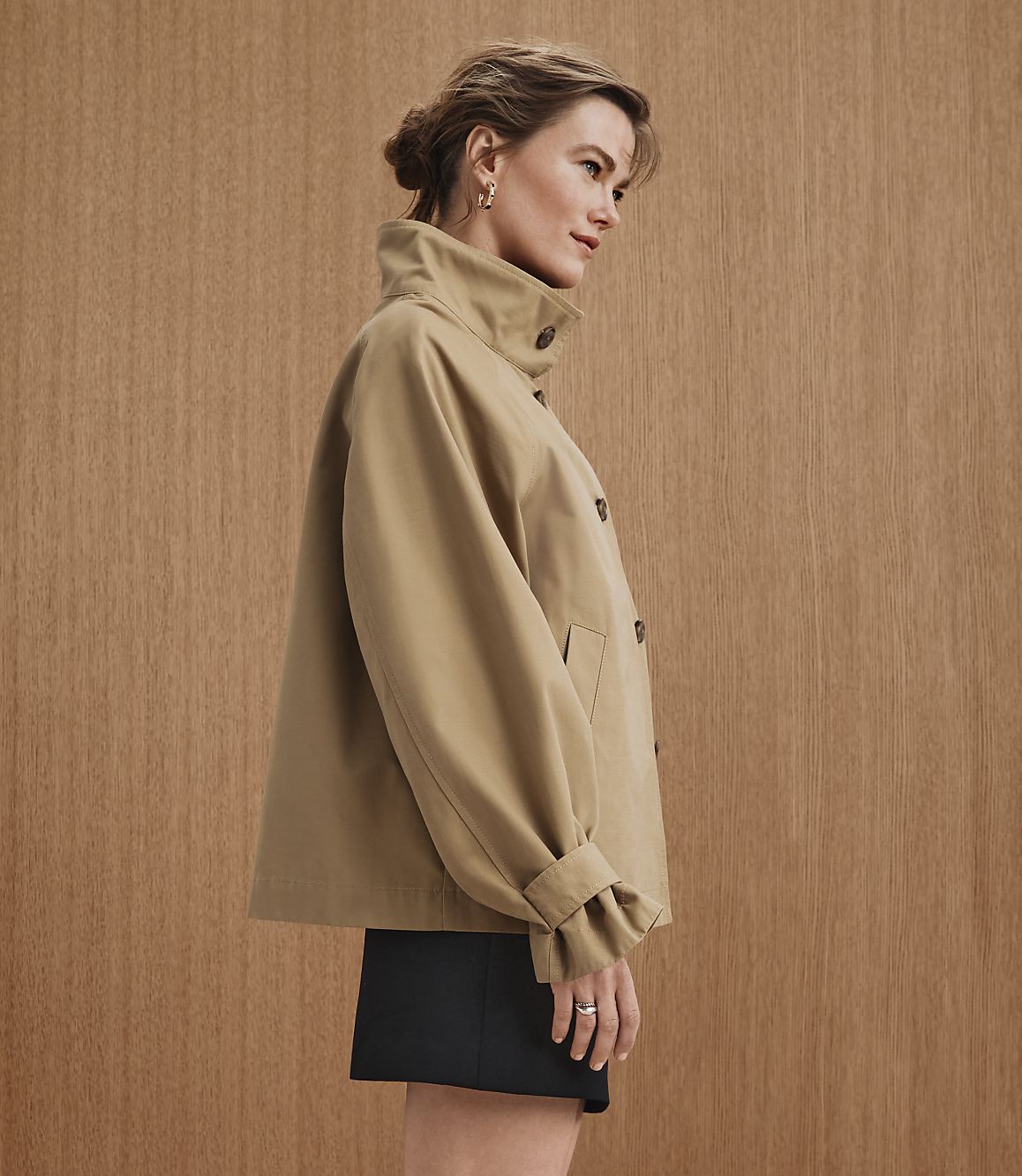 A woman in a tan jacket and black skirt stands against a warm, wood-paneled background.