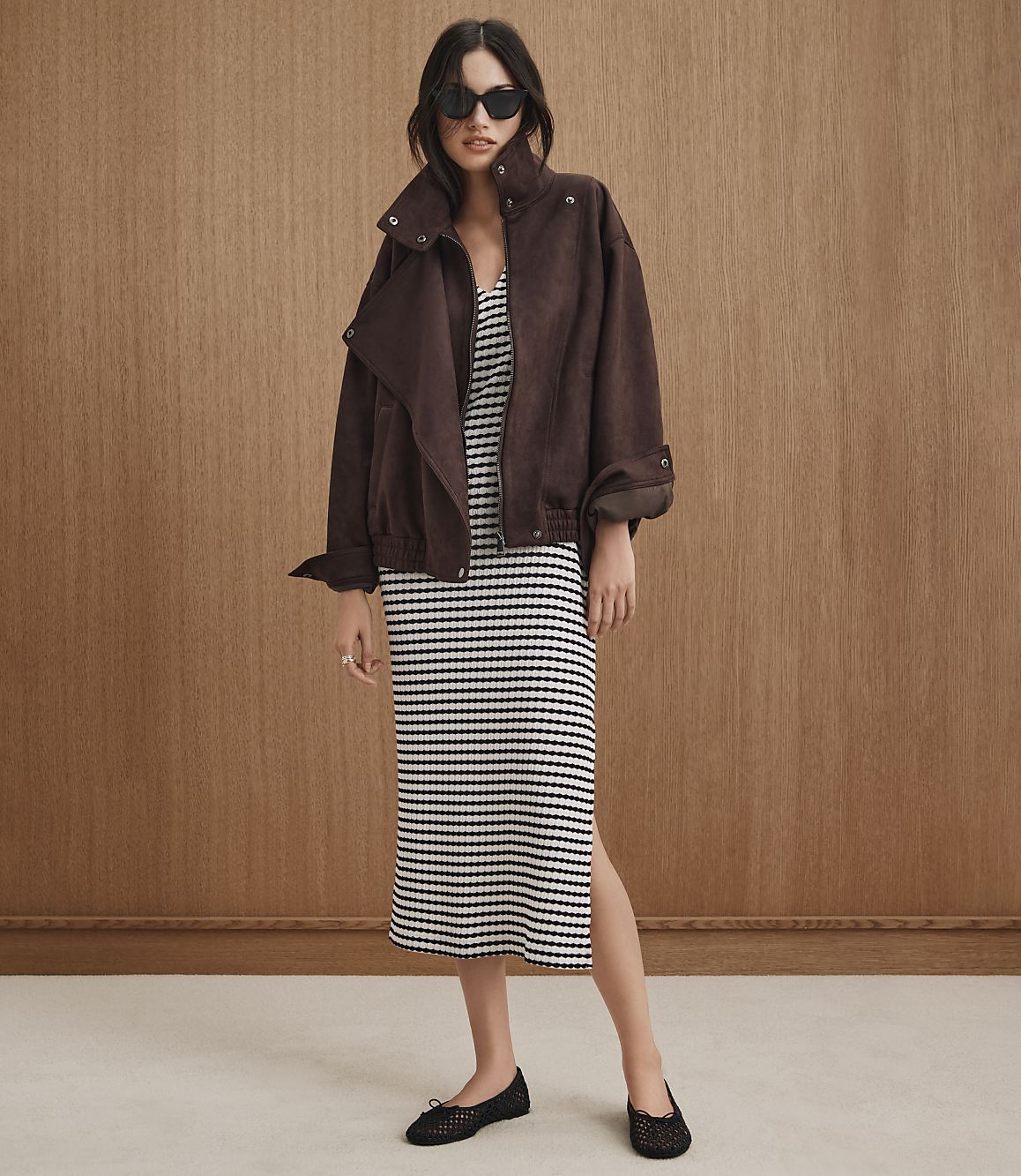 A woman in a brown jacket and striped dress stands against a wood-paneled background.