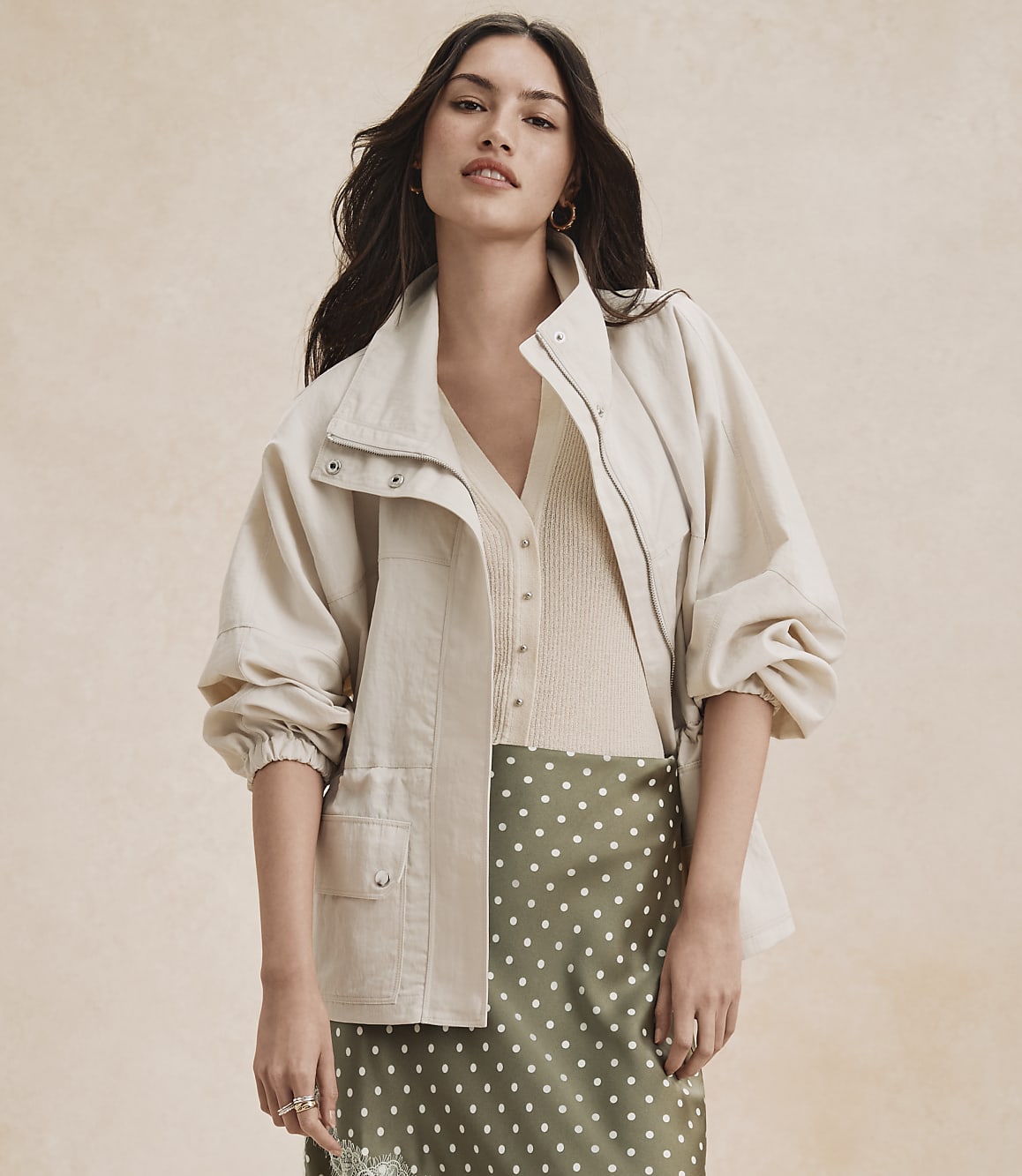A woman in a cream jacket and green polka dot skirt stands against a textured beige background.