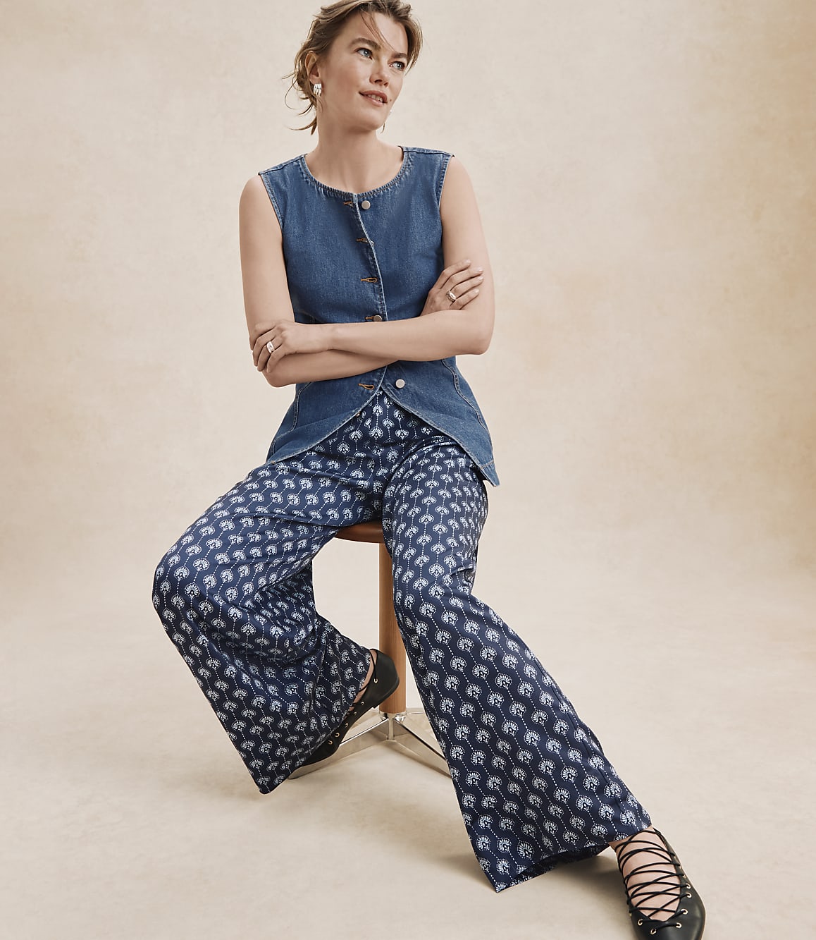A woman in a denim vest and patterned pants sits on a stool against a plain, light-colored background.