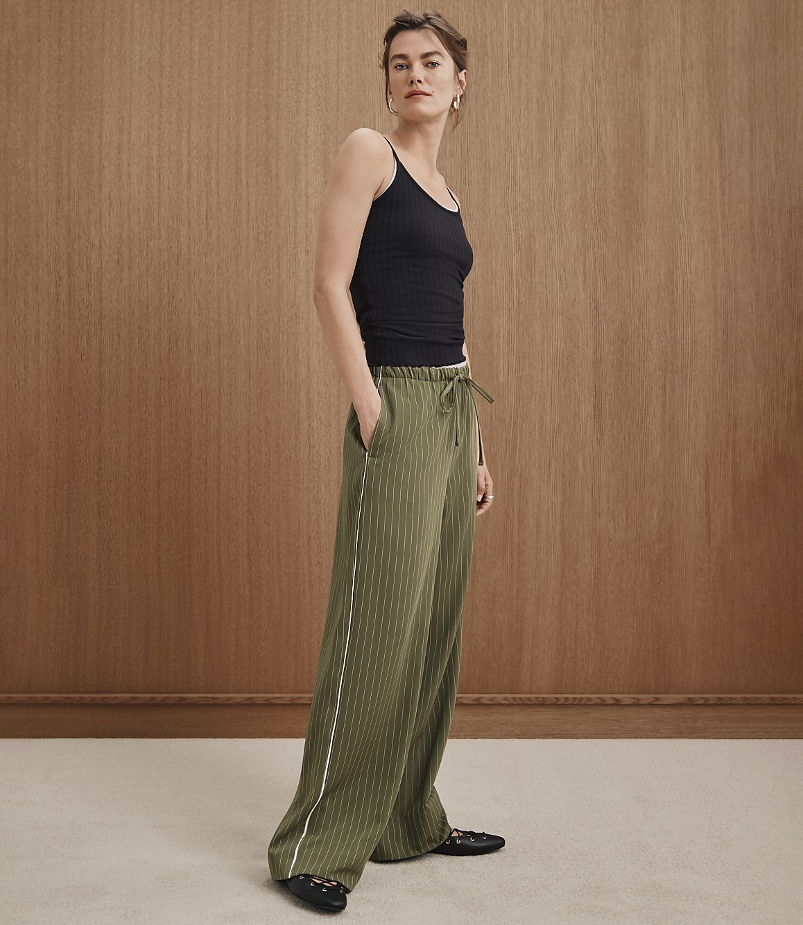 A woman in a black tank top and olive green striped pants stands against a warm, wood-paneled background.