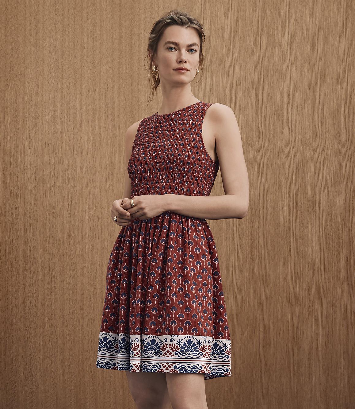 A woman in a patterned red dress stands against a textured brown background.