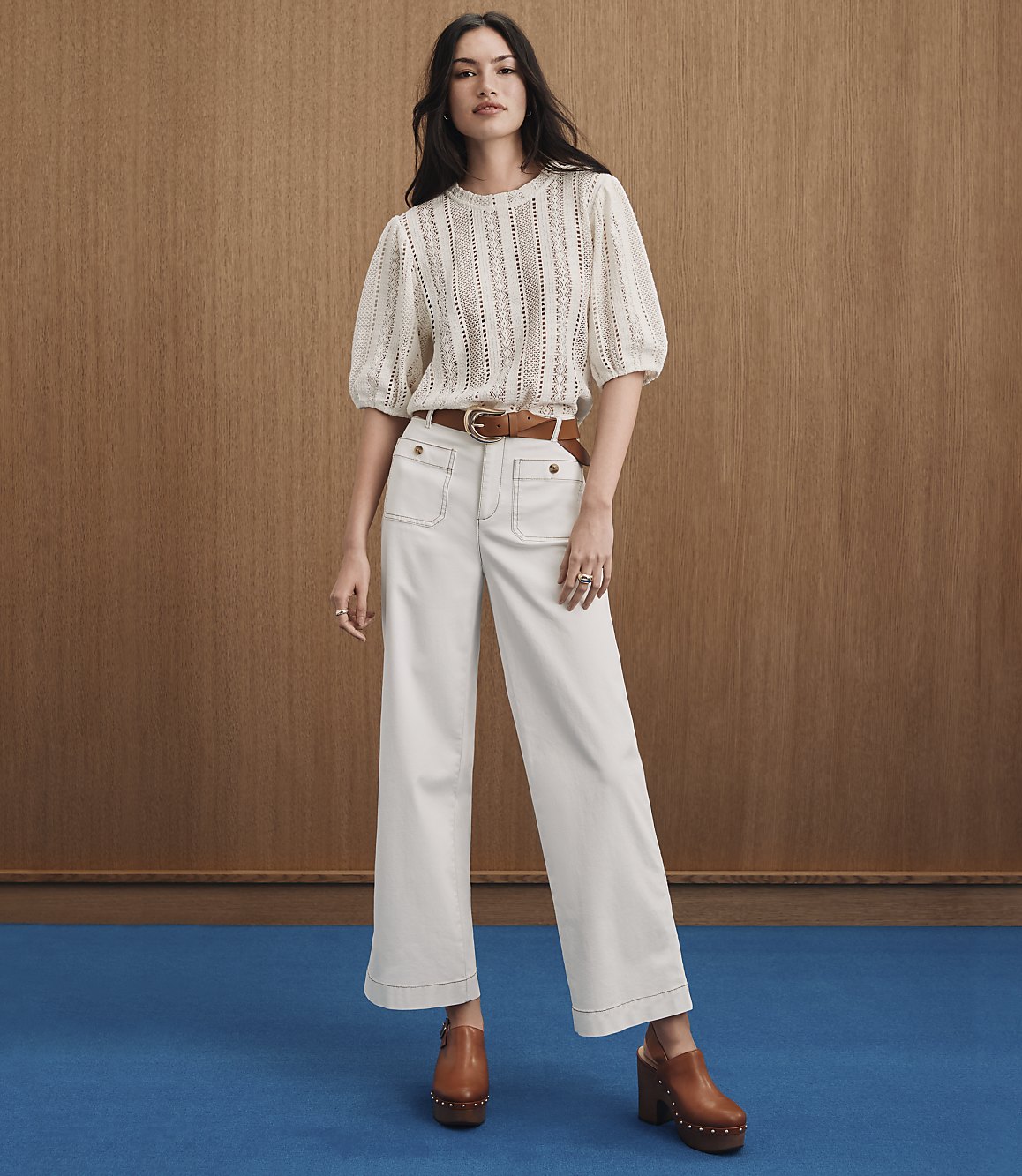 A woman in a cream-colored eyelet top and wide-leg pants stands against a wood-paneled background with a blue floor.