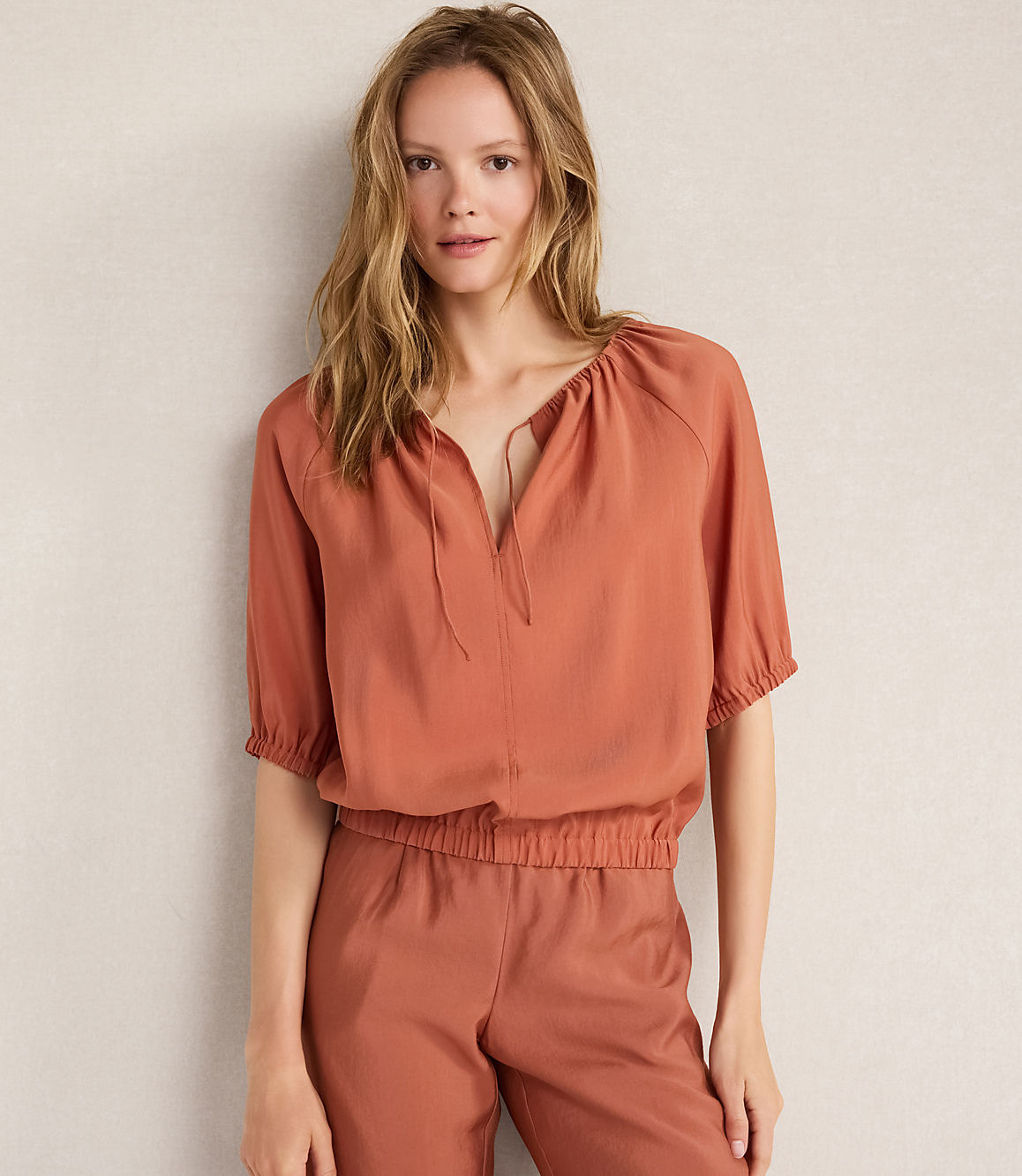 A woman in a rust-colored top and pants stands against a plain, light-colored wall.