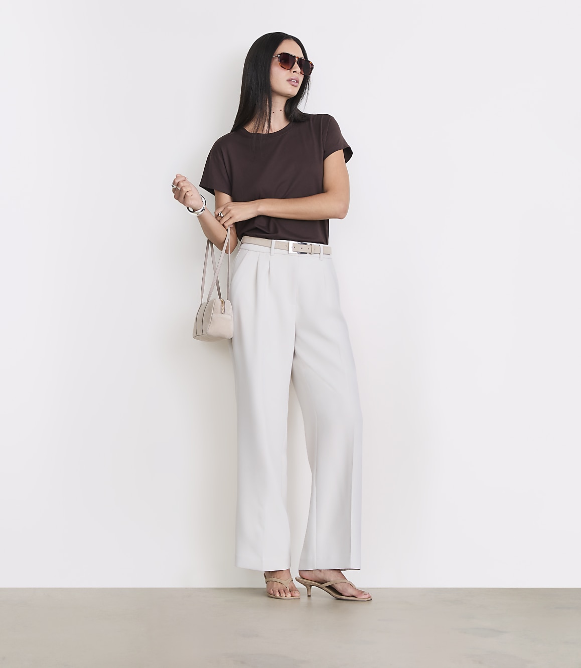 A woman in a brown t-shirt and cream pants stands against a white wall with a light brown floor.