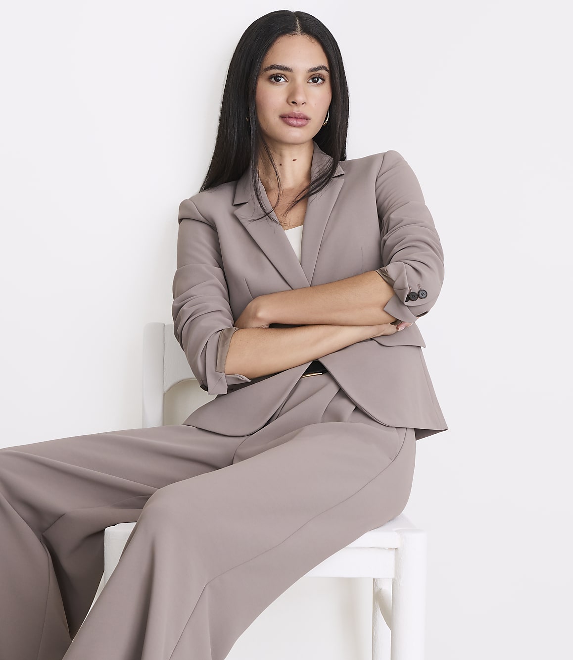 A woman in a taupe pantsuit sits with her arms crossed against a plain white background.