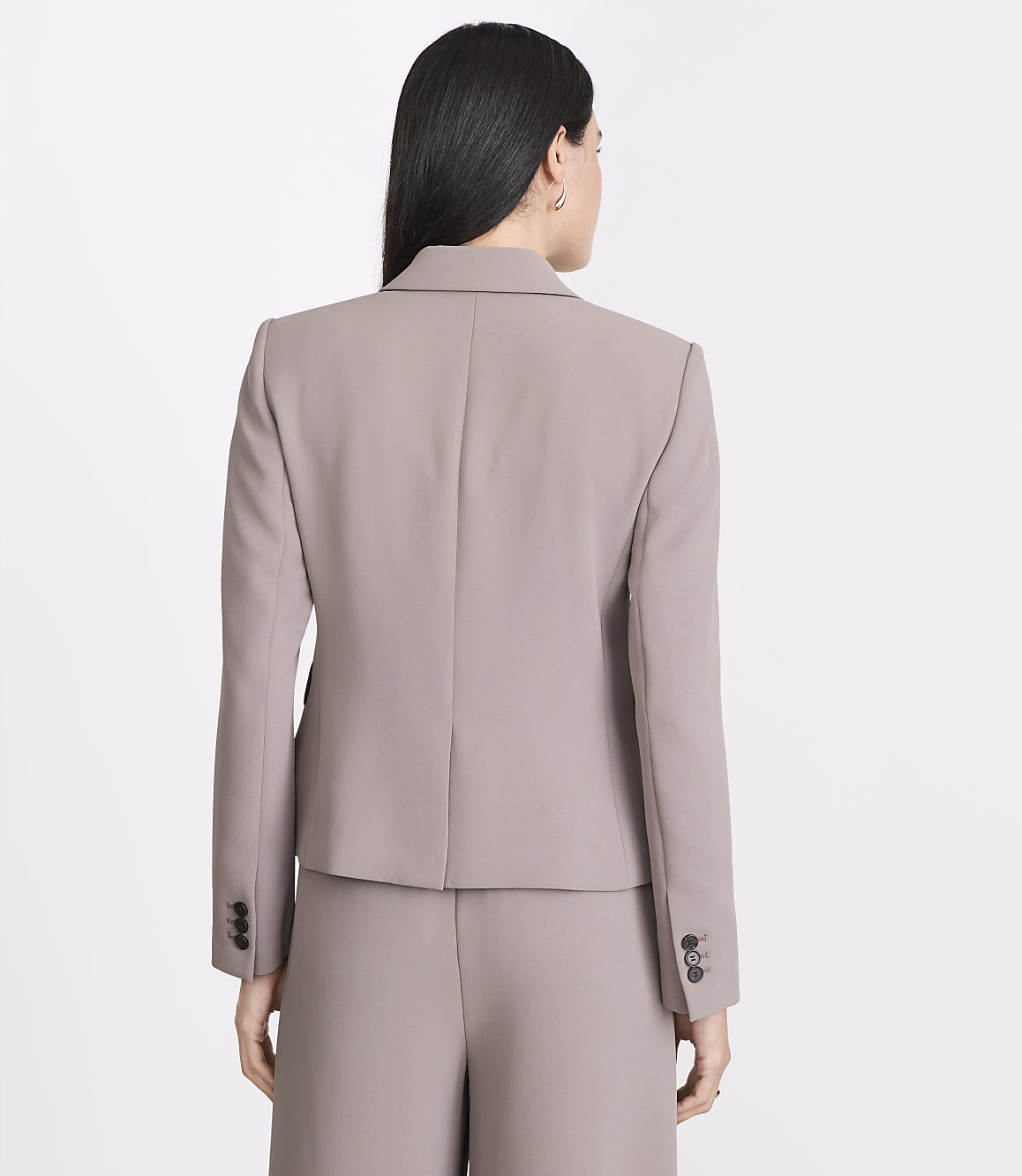 A woman in a taupe blazer and pants stands against a plain white background.