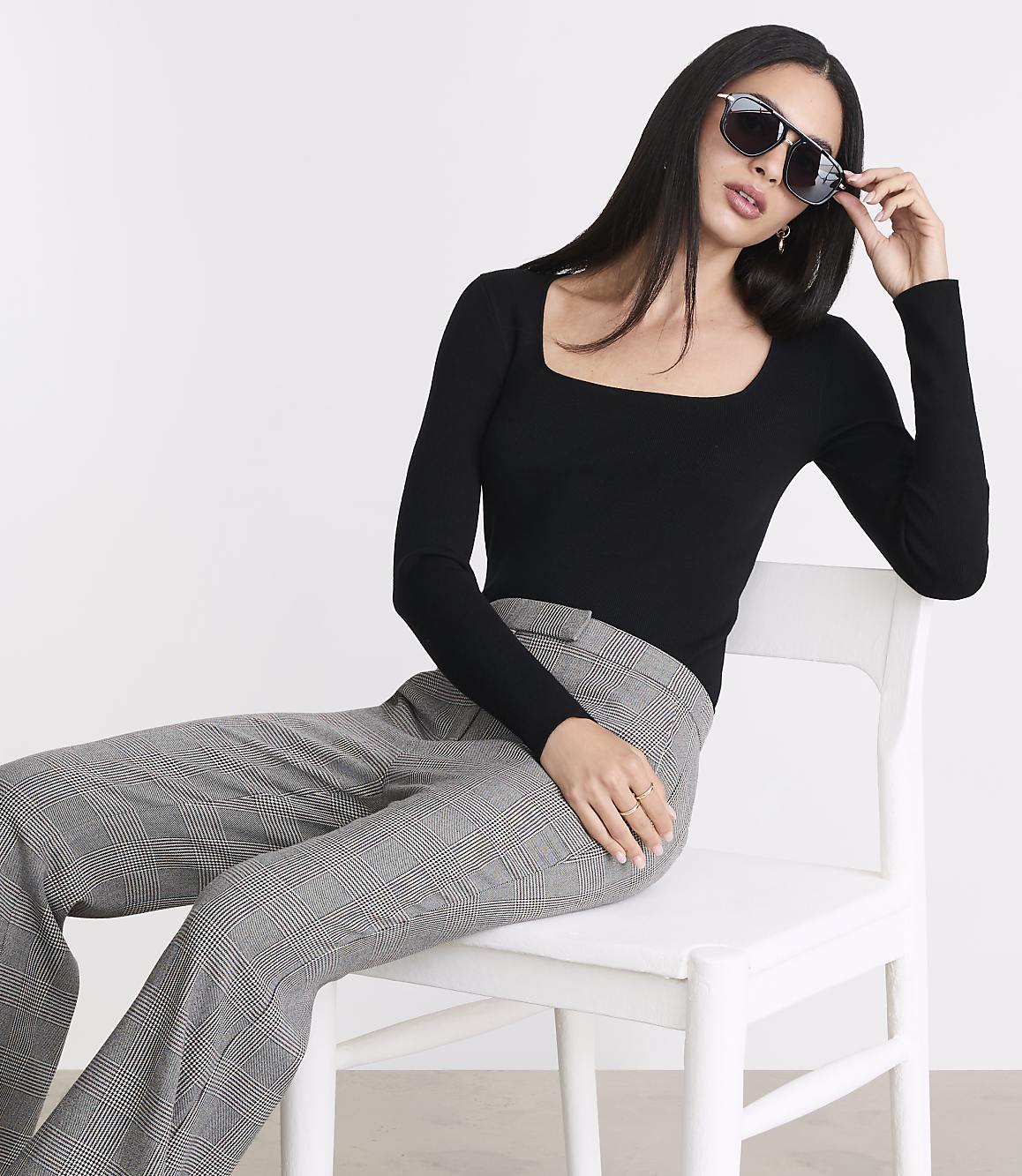 A woman in a black top and plaid pants sits on a white chair against a plain white background, adjusting her sunglasses.