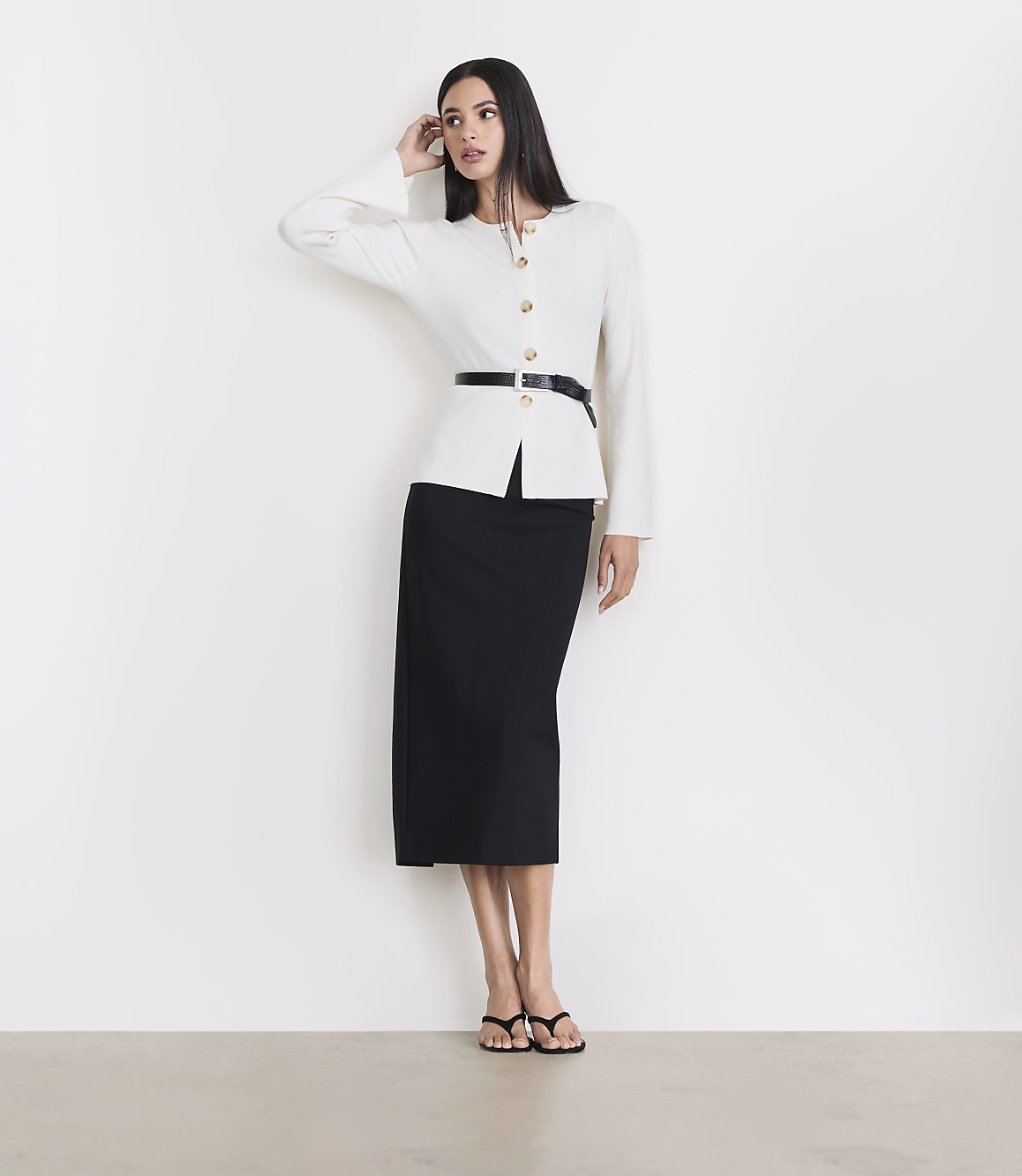 A woman in a white jacket and black skirt stands against a white wall with a light gray floor in the foreground.
