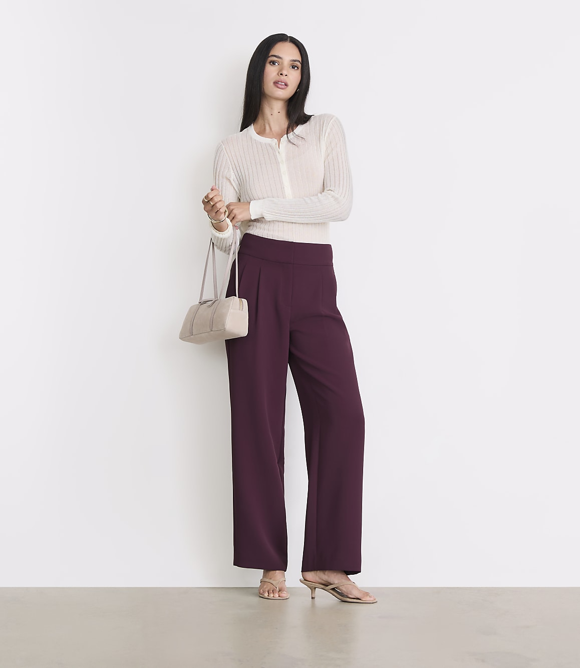 A woman in a cream cardigan and plum wide-leg pants stands against a white background, holding a cream handbag.