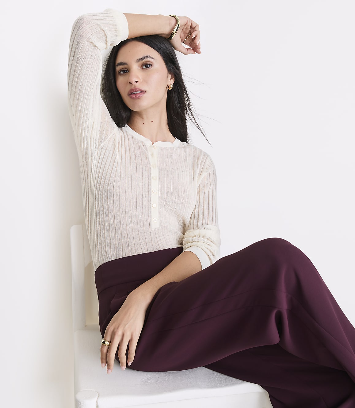A woman in a cream-colored ribbed shirt and burgundy pants sits on a white chair against a plain white background.