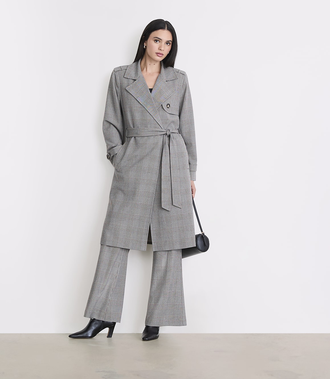 A woman in a plaid trench coat and matching pants stands against a plain white background, holding a black purse.