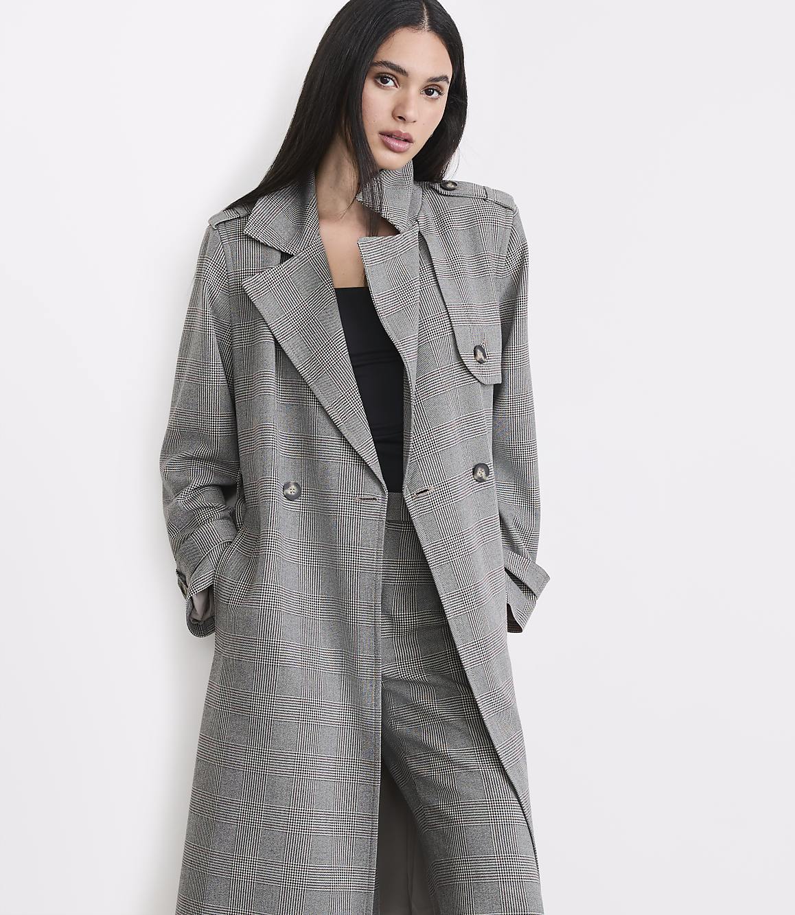 A woman in a plaid trench coat and pants stands against a plain white background.
