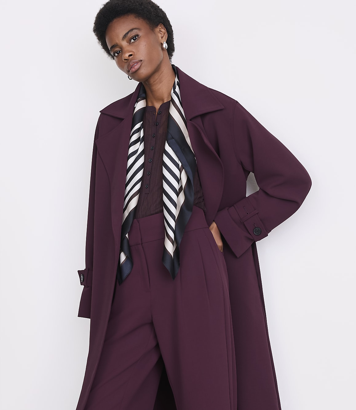 A woman in a burgundy trench coat and matching pants stands against a plain white background.