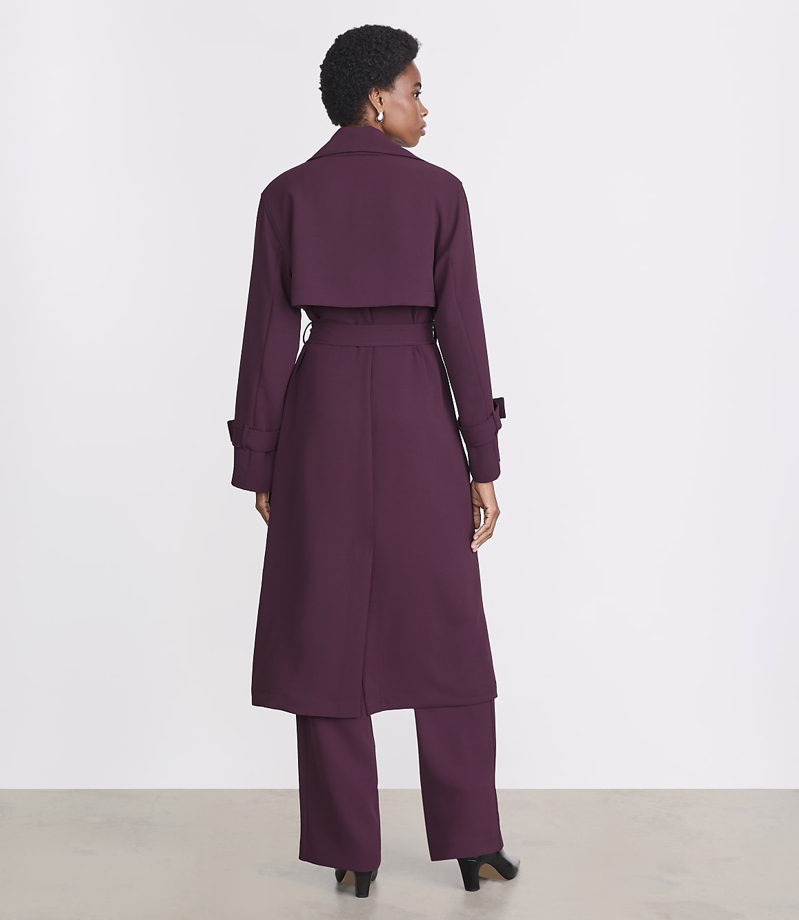 A woman in a plum trench coat and matching pants stands against a plain white background.