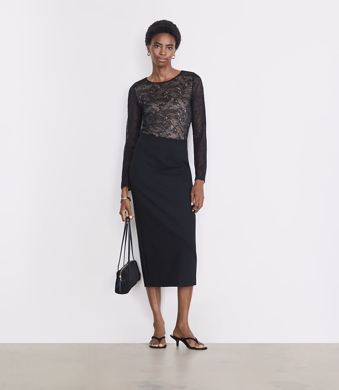 A woman in a black lace top and pencil skirt stands against a plain white background, holding a small black purse.