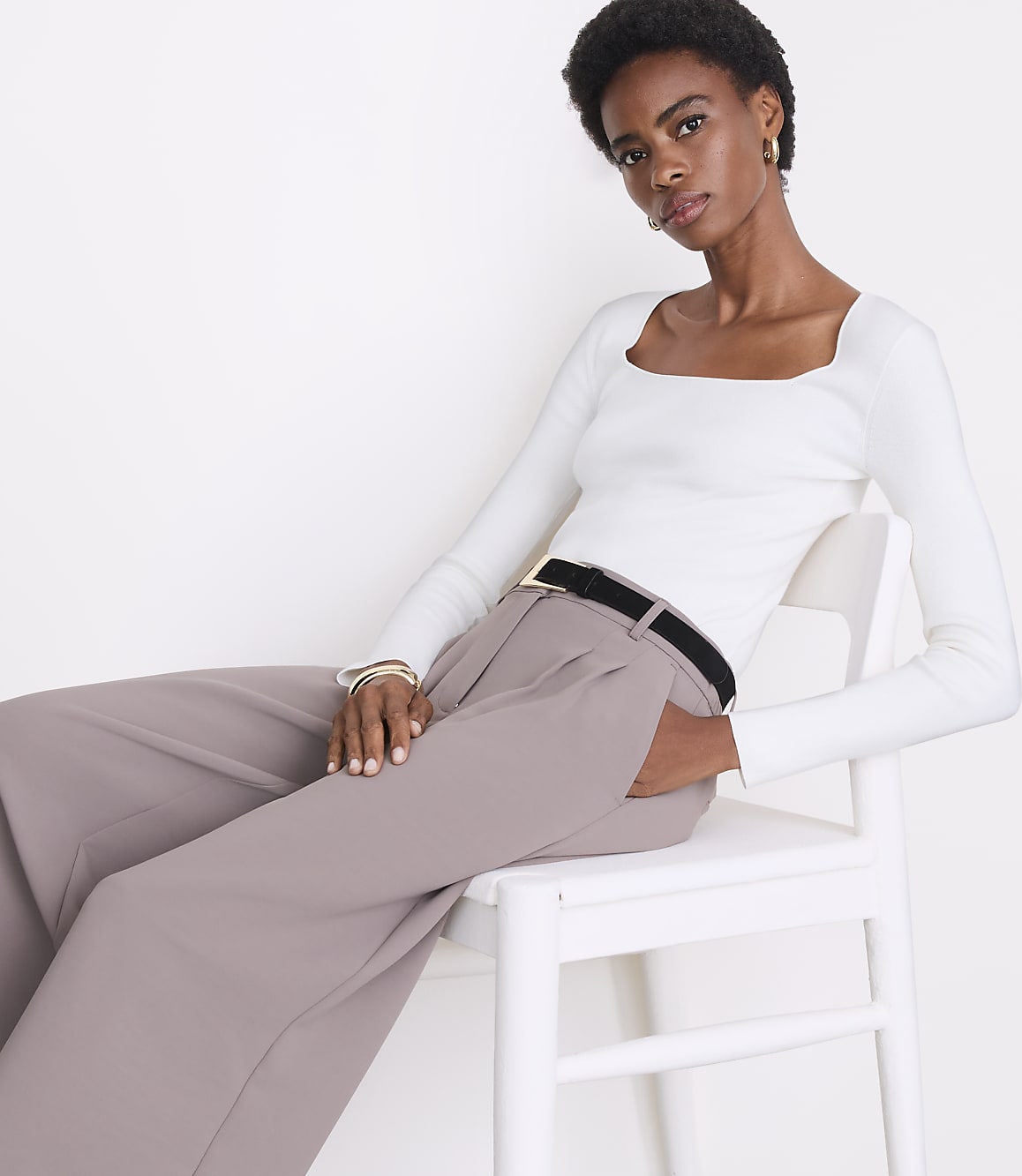 A woman in a white top and taupe pants sits on a white chair against a plain white background.