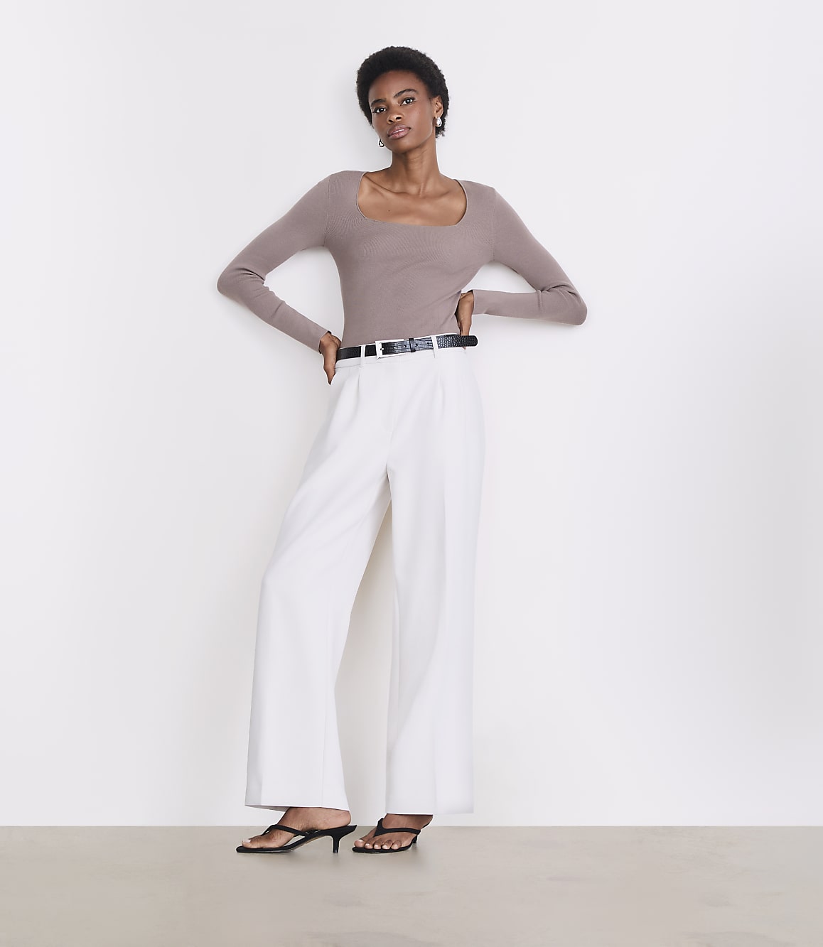 A woman in a taupe square-neck top and white wide-leg pants stands against a plain white background.