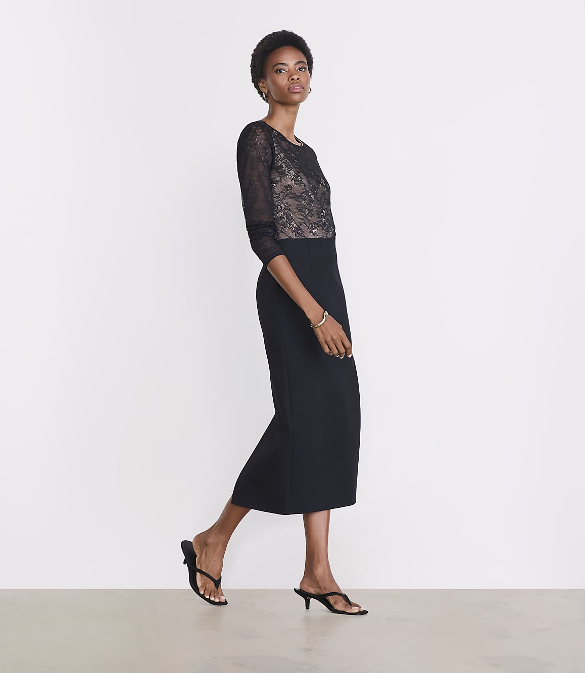 A woman in a black lace-bodice dress and heeled sandals stands against a plain white background.