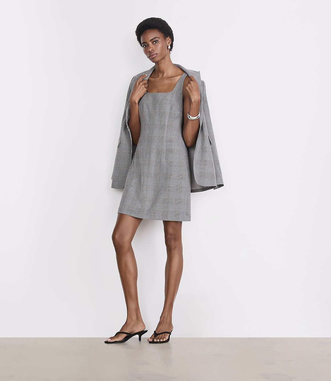 A woman in a plaid dress and heels stands against a plain white background with a blazer draped over her shoulders.